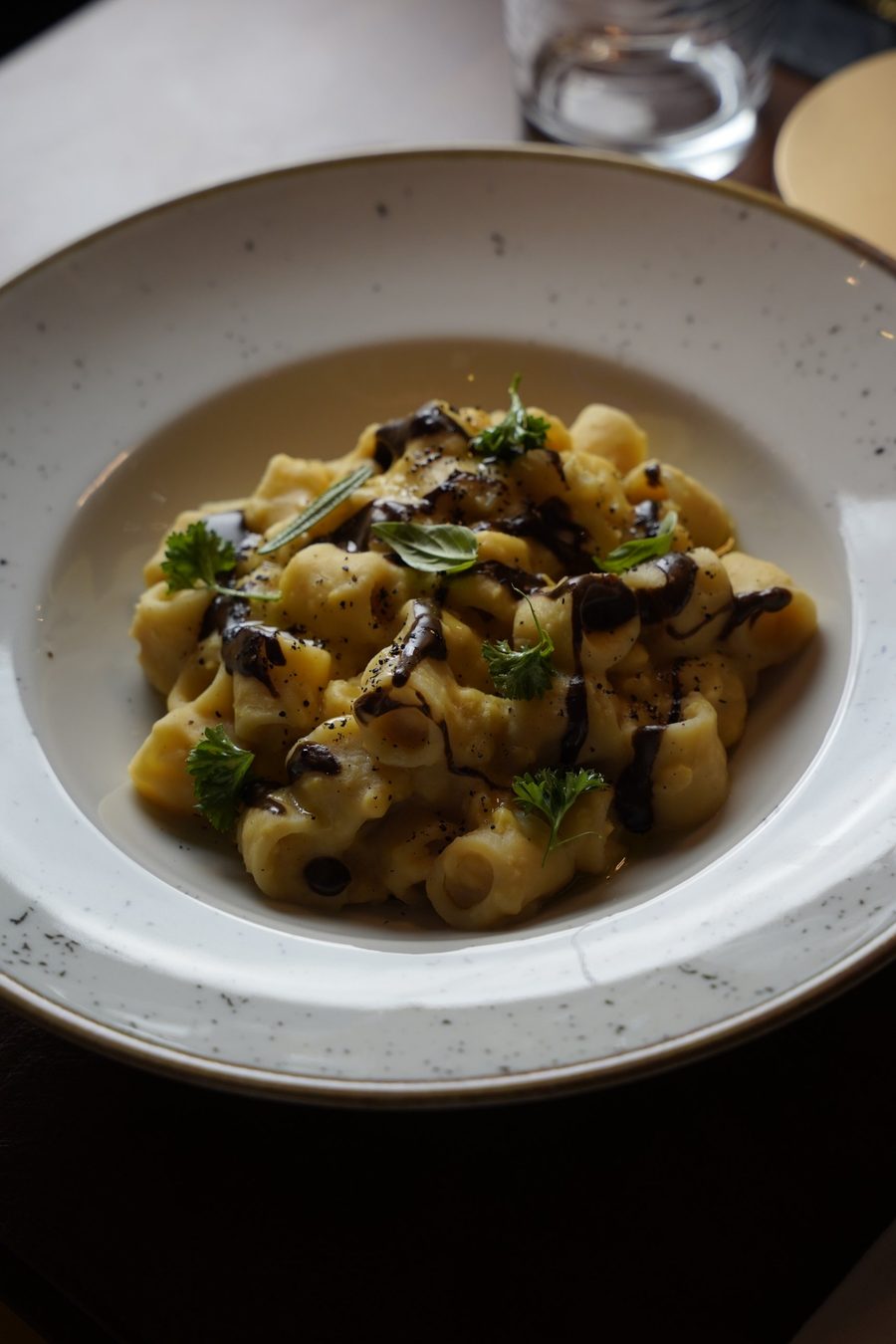 Pasta corta in crema gialla con filo di salsa scura, prezzemolo e basilico, in un piatto fondo puntinato