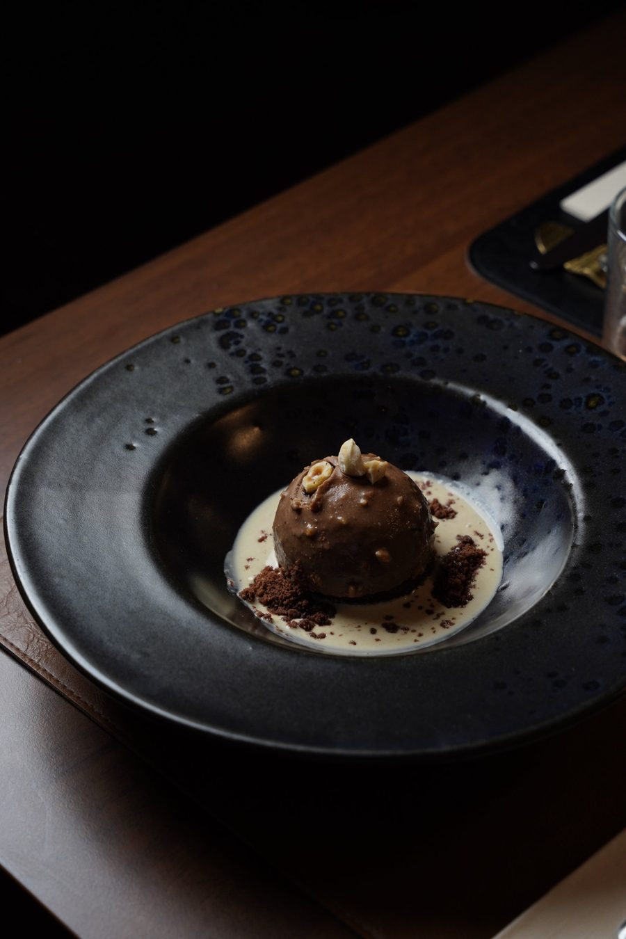 Cupola al cioccolato con nocciole su crema chiara e crumble, in un piatto nero smaltato
