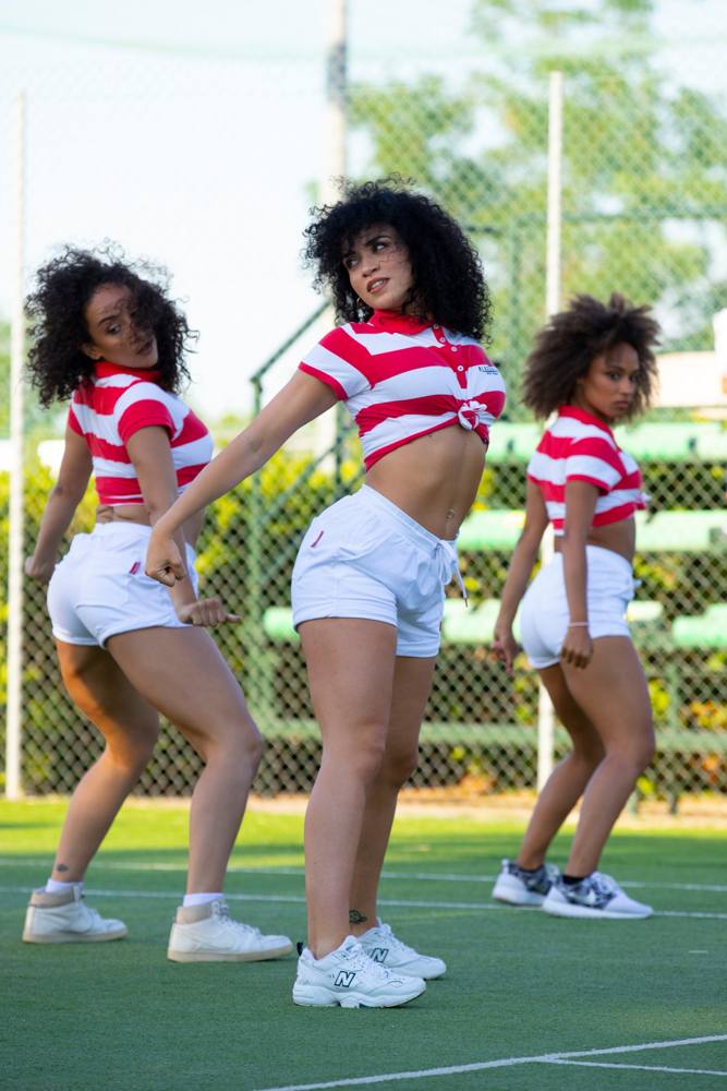 Tre ballerine in polo a righe rosse e bianche e pantaloncini bianchi, girate a guardare sopra la spalla su un campo da tennis