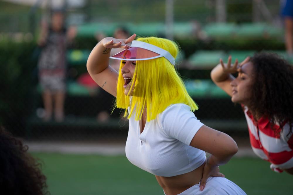 La donna con il caschetto giallo si porta una mano alla visiera, accanto a una ballerina dai capelli ricci