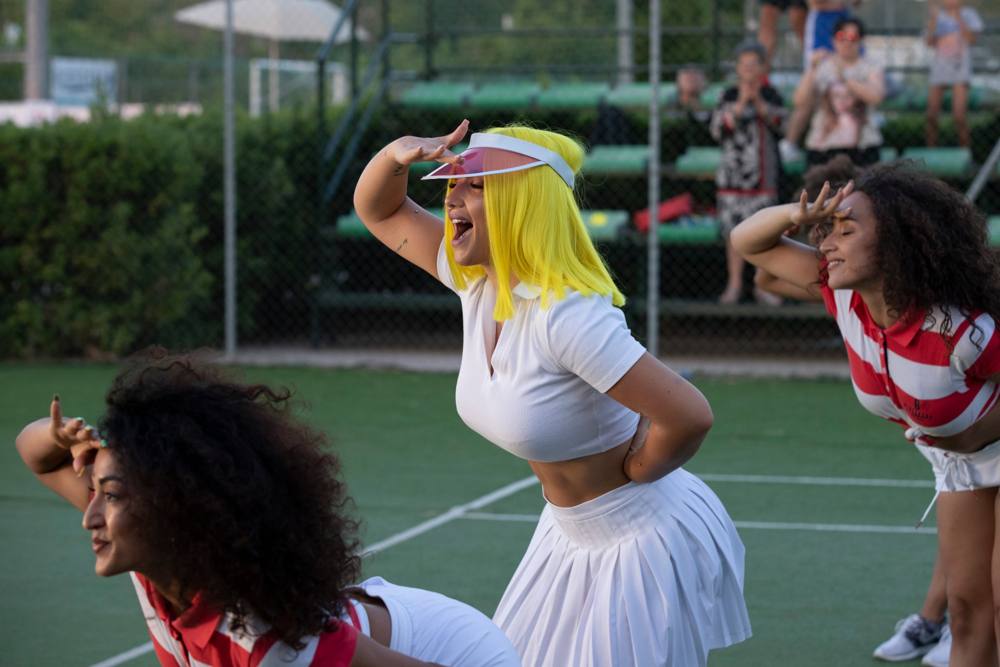 La donna con il caschetto giallo si ripara gli occhi con la mano mentre canta, tra due ballerine