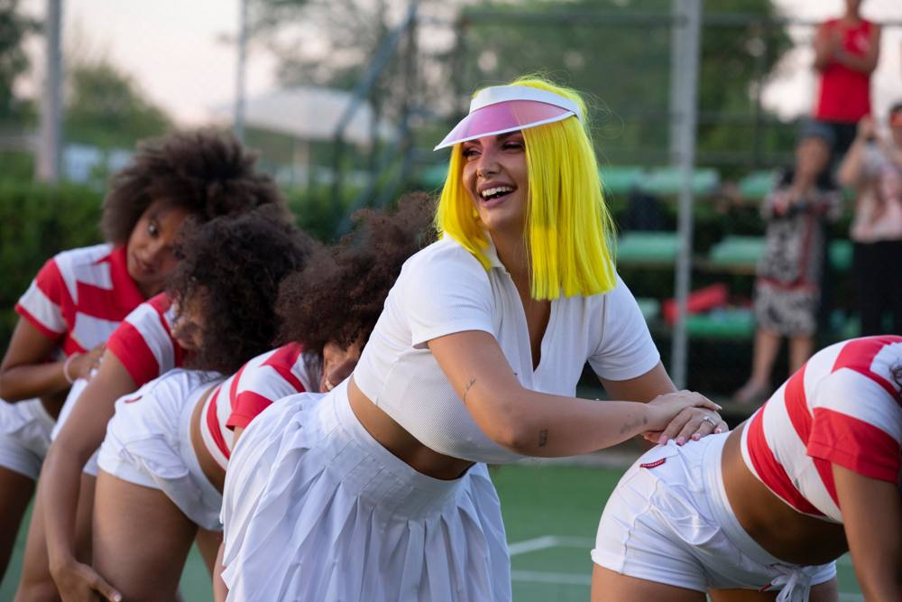La donna con il caschetto giallo ride a testa alta durante una coreografia, con le ballerine chinate accanto a lei