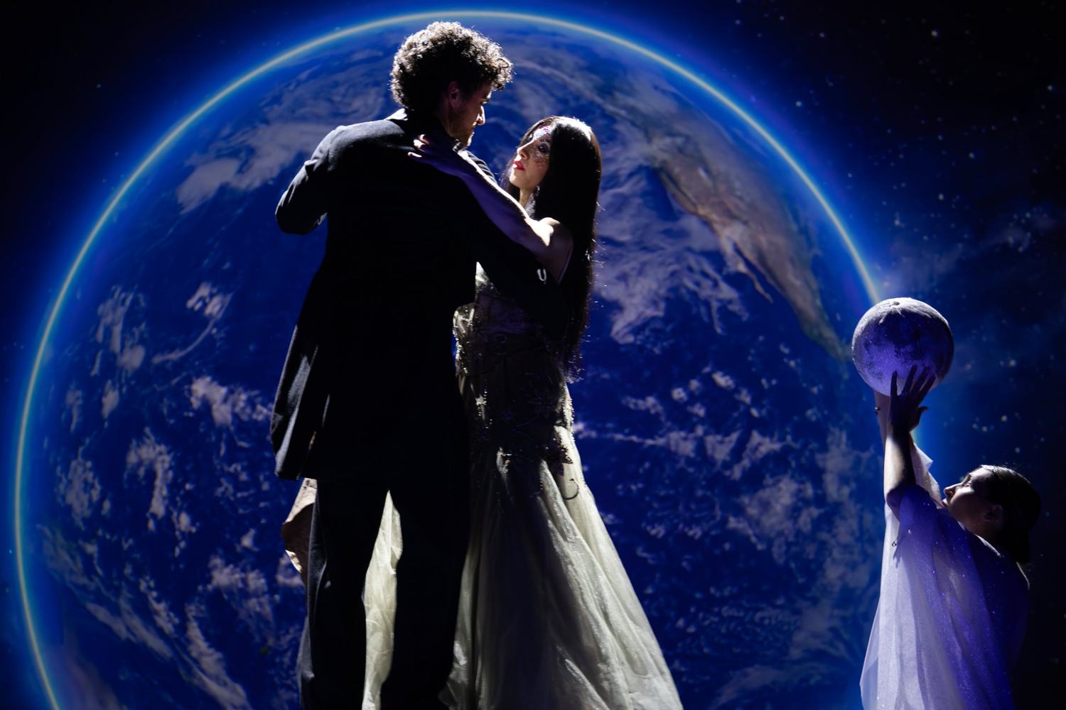 Un uomo e una donna in abito lungo ballano in controluce davanti a un enorme pianeta Terra; a destra una figura in bianco solleva una piccola luna