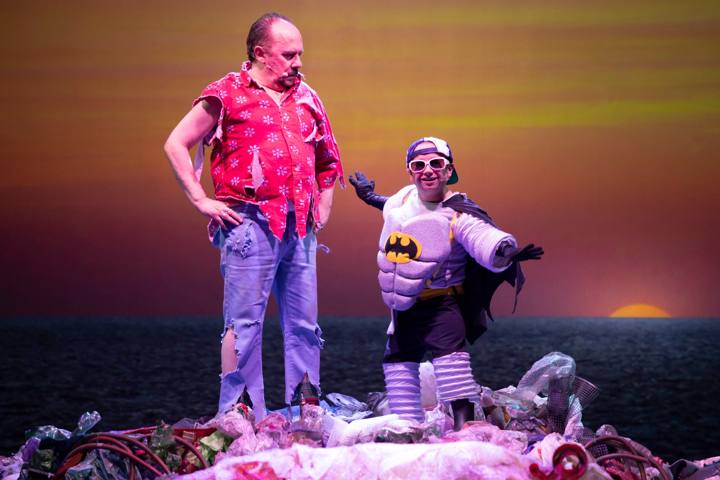 Un uomo con camicia rossa e jeans strappati accanto a una figura con costume da pipistrello e occhiali bianchi, sul tramonto sul mare