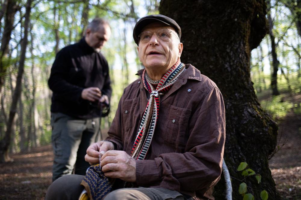 Un uomo anziano con coppola e sciarpa a righe cuce con ago e filo nel bosco