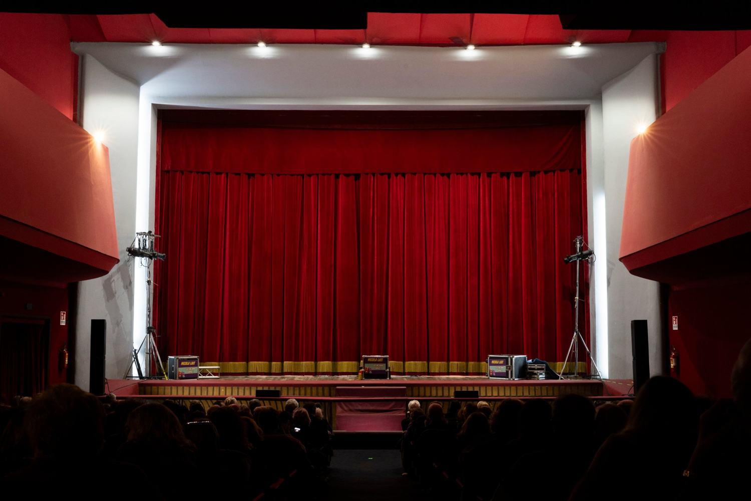 La sala del teatro vista dalla platea: il grande sipario rosso chiuso, due valigie da tour sul palco e il pubblico in ombra