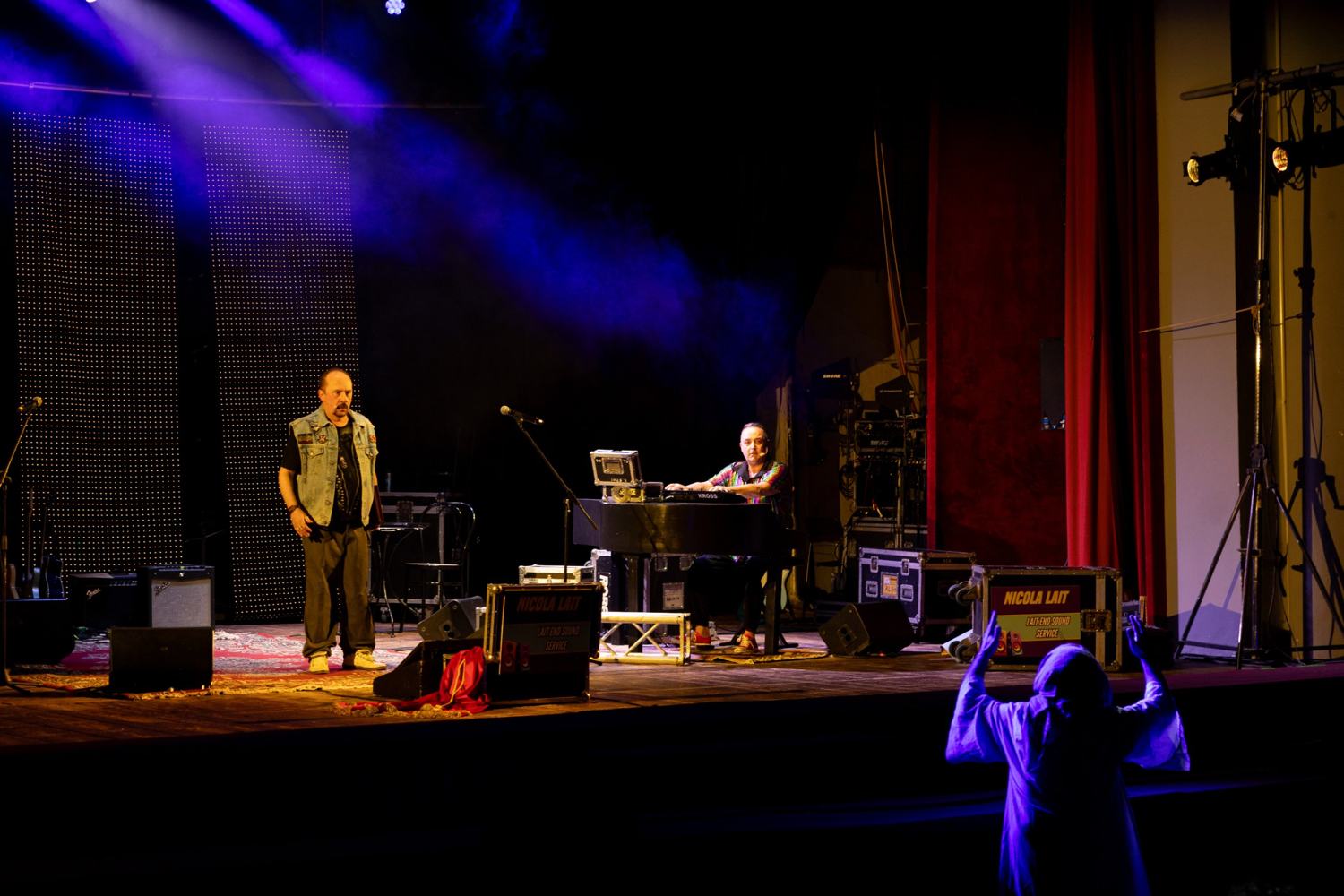 Palco con luci viola nel fumo: un uomo in gilet di jeans in piedi, un tastierista seduto al pianoforte e, in primo piano, una figura con le braccia alzate