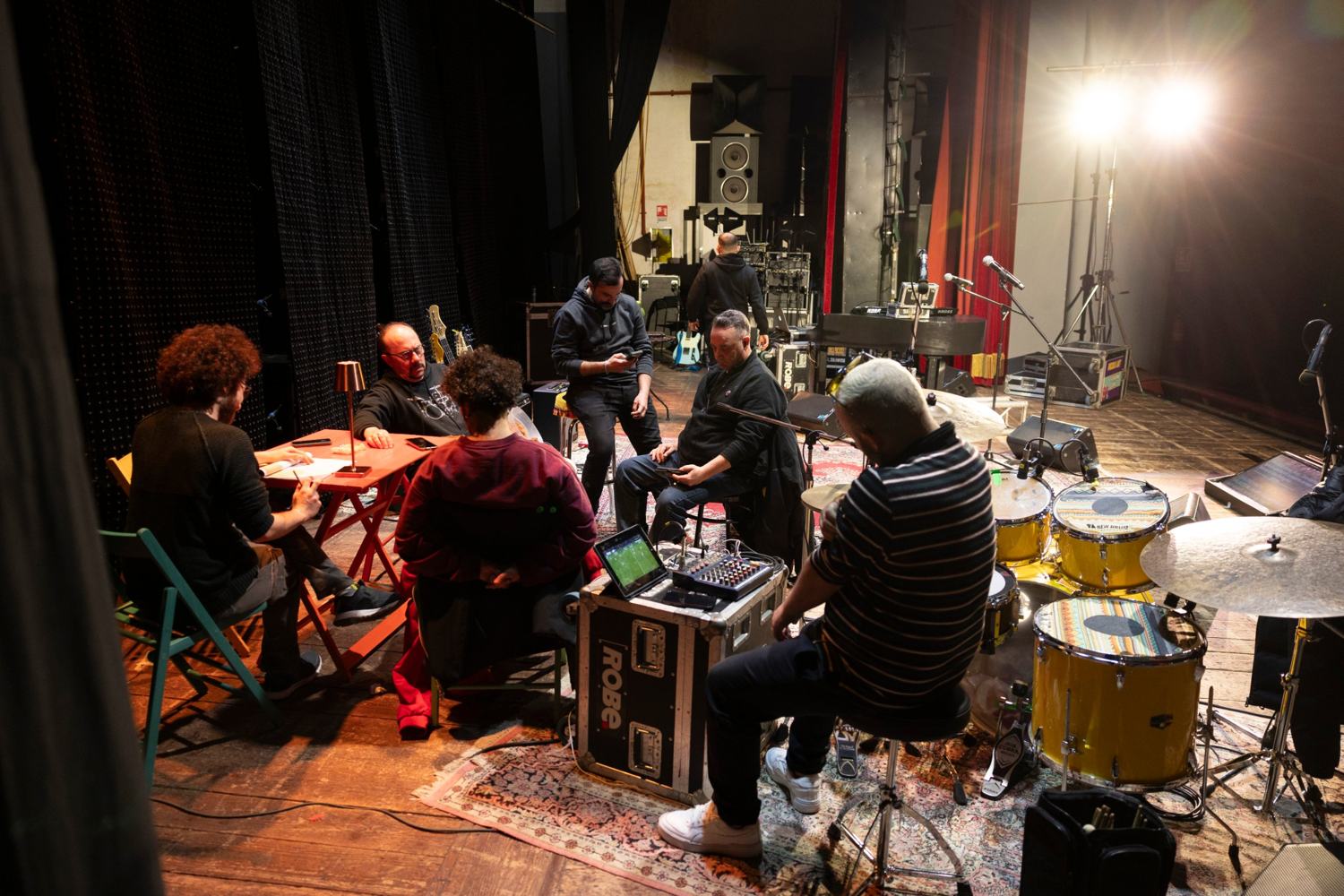 Palco durante le prove: alcune persone attorno a un tavolino rosso, una batteria gialla, una valigia da tour con un mixer e un faro acceso sullo sfondo