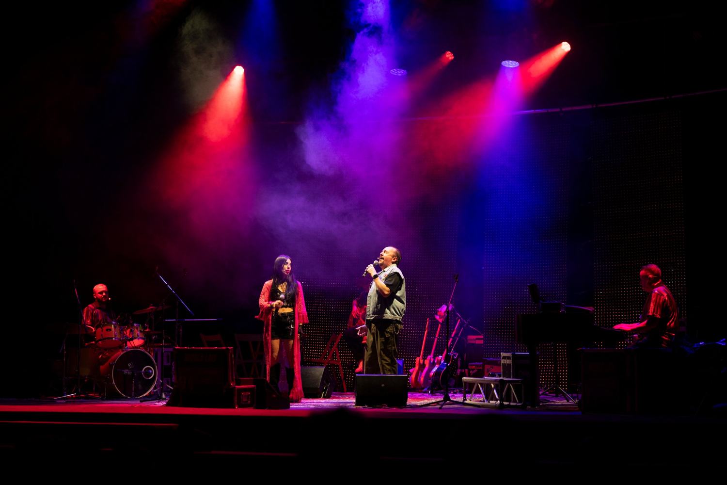 Sul palco, con fumo e fasci di luce rossi e blu, una donna con lunga veste rosa e un uomo con gilet di jeans cantano; ai lati un batterista e un tastierista