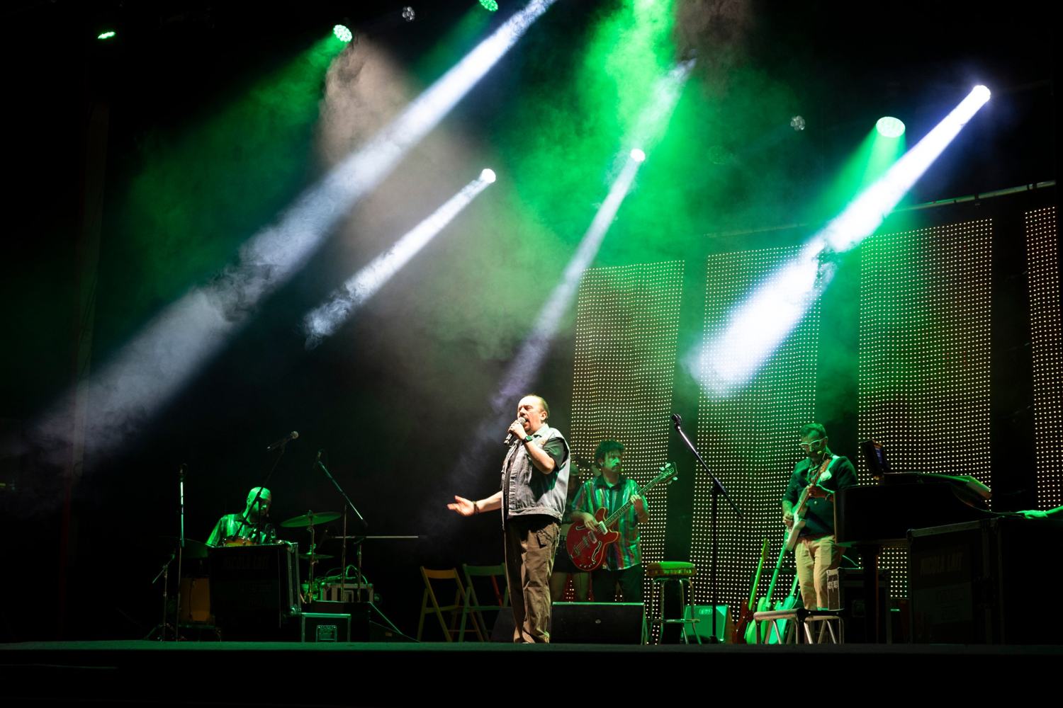 Palco inondato di luci verdi e bianche con fumo: un cantante con gilet di jeans al centro, un bassista e un chitarrista ai lati e un batterista a sinistra